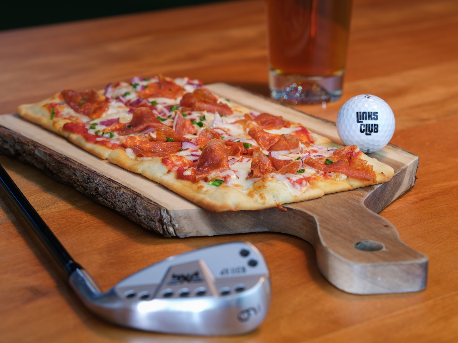 Pepperoni flatbread on a wood board with a Links Club golf ball and a craft beer